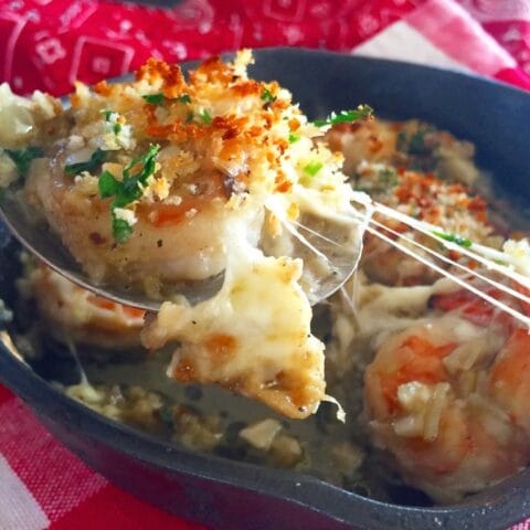 The image shows a serving of baked shrimp with a breadcrumb topping being scooped from a skillet. The shrimp is topped with melted cheese, fresh herbs, and crispy breadcrumbs, making for a delicious, golden-brown presentation. The dish sits on a red and white checkered cloth.