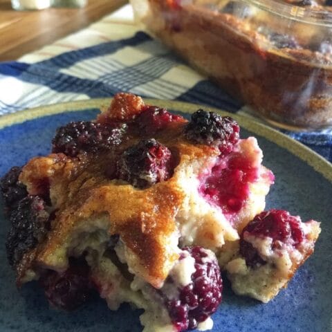 The image shows a serving of blackberry cobbler on a blue plate, with a golden-brown crust and juicy blackberries. In the background, a glass baking dish with the remaining cobbler can be seen. The setting is casual, likely indicating a freshly baked dessert ready to be enjoyed.