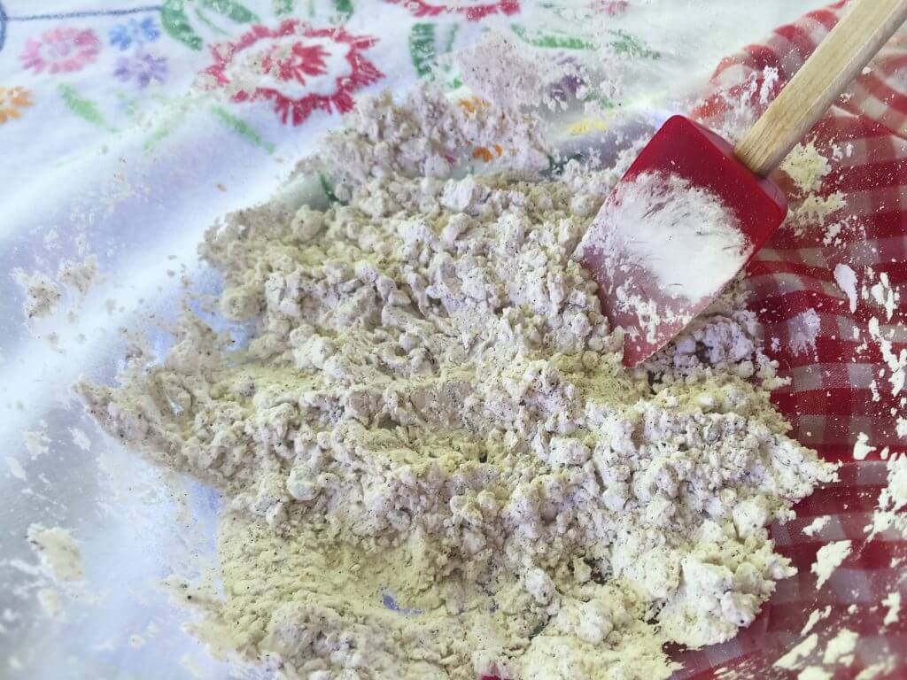 A mixing bowl containing a lumpy flour mixture with a red spatula is set on a table covered with a floral-patterned tablecloth and a red and white checkered cloth.