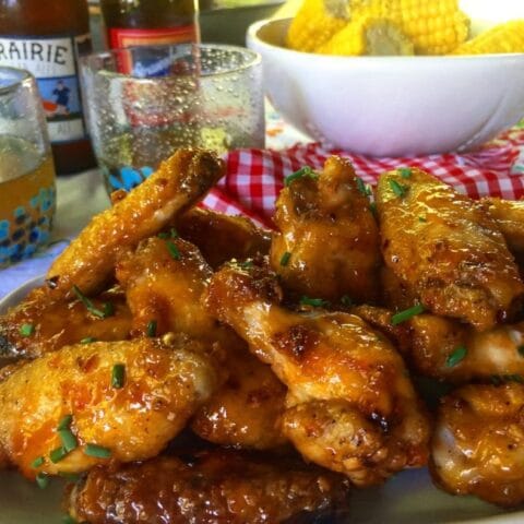The image shows a plate of golden, glazed chicken wings garnished with herbs. In the background, there is a bowl of corn on the cob and bottles of beer, suggesting a casual, outdoor meal setting. The scene evokes a relaxed and tasty gathering.