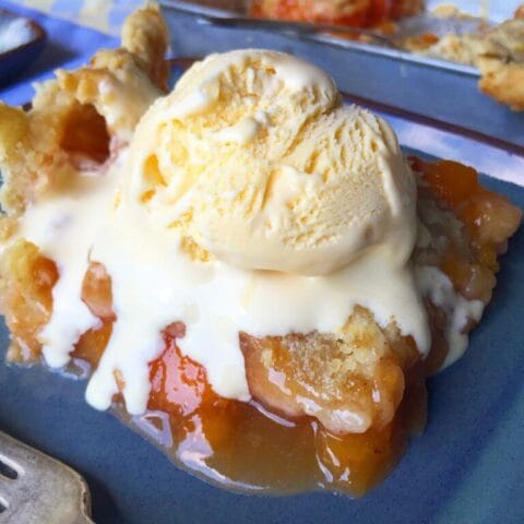 A slice of peach pie topped with a scoop of melting vanilla ice cream on a blue plate.