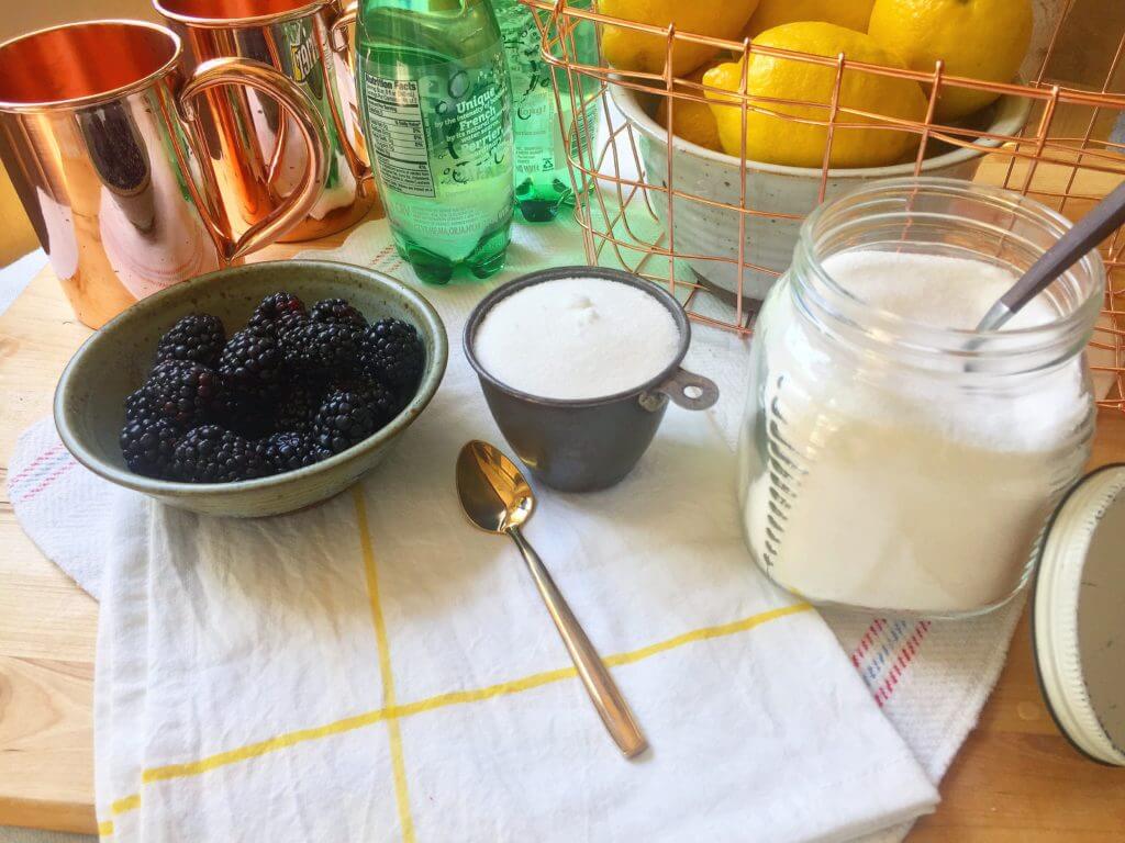 A bowl of blackberries, a cup of sugar, a jar of sugar, and a spoon on a cloth with lemons, copper mugs, and bottles in the background.