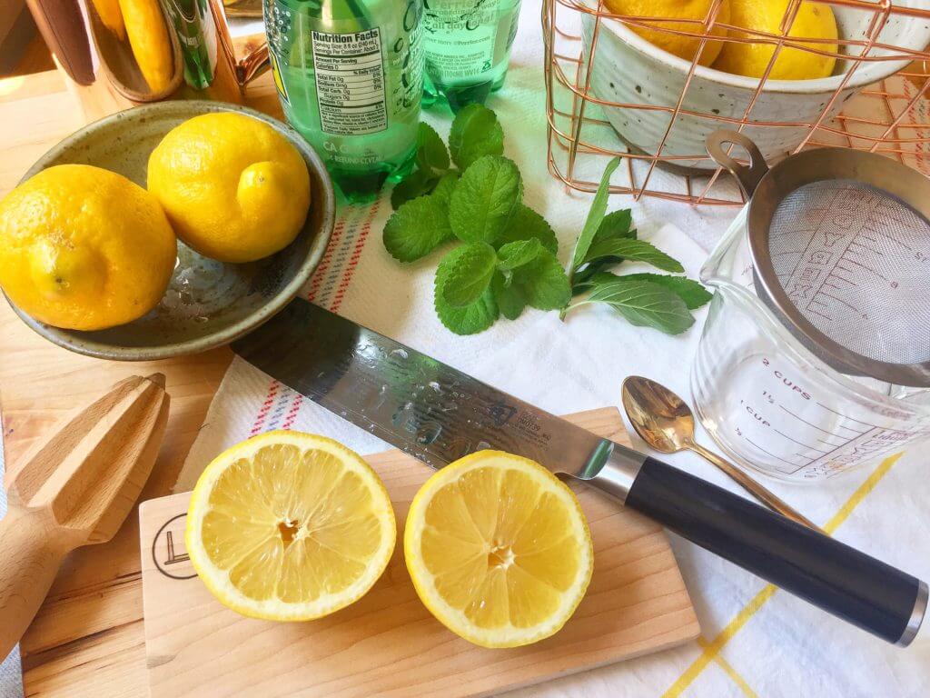 Whole and halved lemons with a knife, mint leaves, a citrus reamer, and a measuring cup on a kitchen counter.