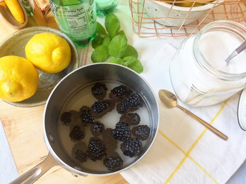 A saucepan with blackberries and water on a kitchen counter, surrounded by lemons, mint leaves, a jar of sugar, and a spoon.