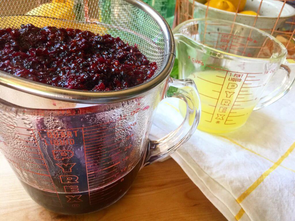 Two measuring cups, one filled with strained blackberry mixture and the other with lemon juice, on a kitchen counter.