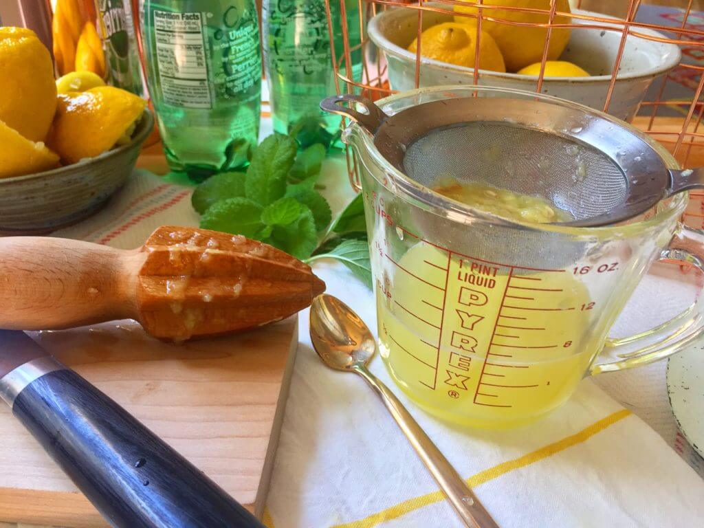 A measuring cup of strained lemon juice with a citrus reamer, lemons, mint leaves, and a spoon on a kitchen counter.