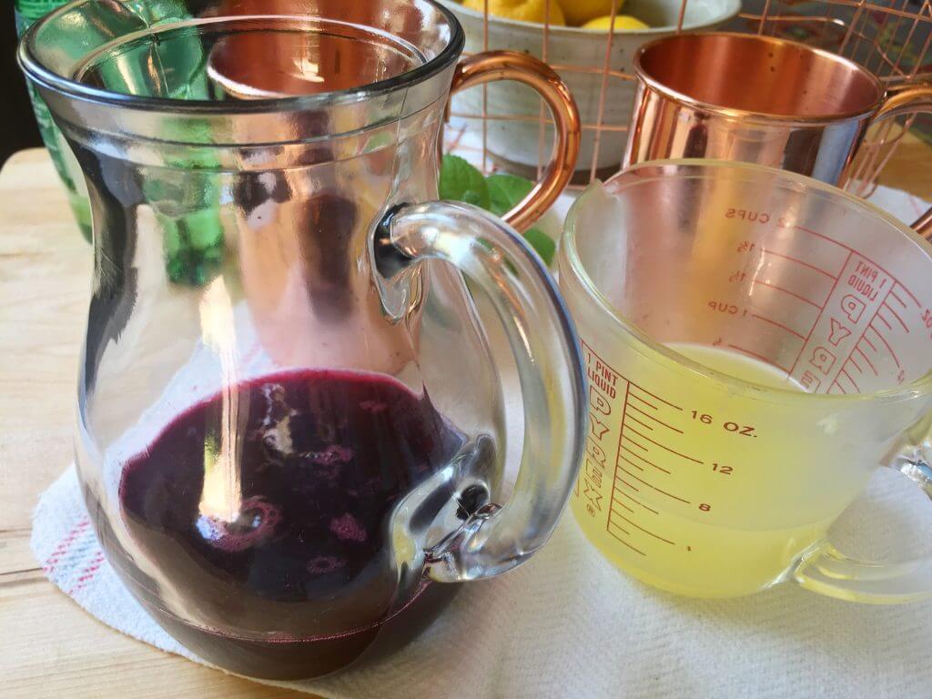 A glass pitcher with a red beverage and a measuring cup with lemon juice on a kitchen counter, with copper mugs and lemons in the background.