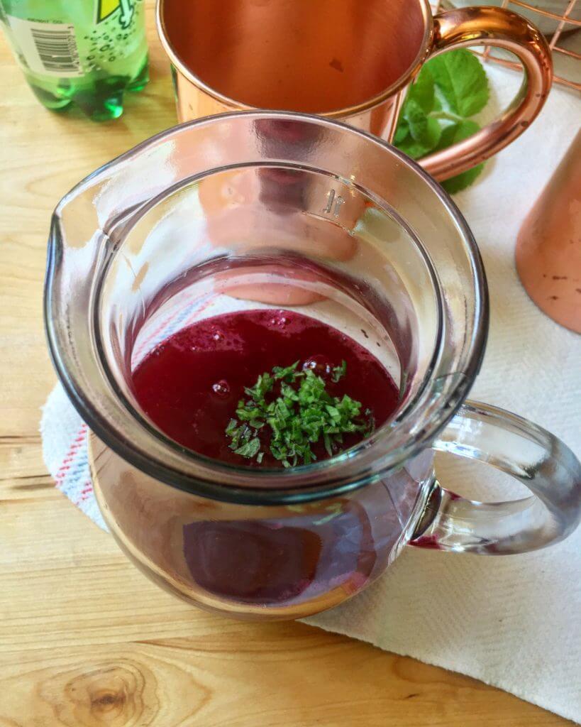 A glass pitcher filled with a red beverage topped with chopped mint, with copper mugs and a bottle in the background.
