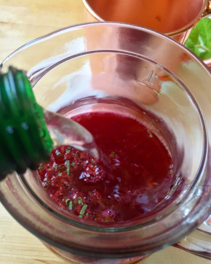 Pouring liquid from a green bottle into a glass pitcher containing a red beverage with chopped mint.