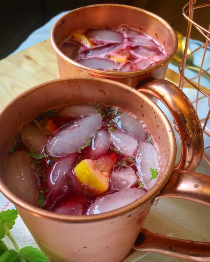 Two copper mugs filled with a red beverage, ice, lemon slices, and mint on a kitchen counter.