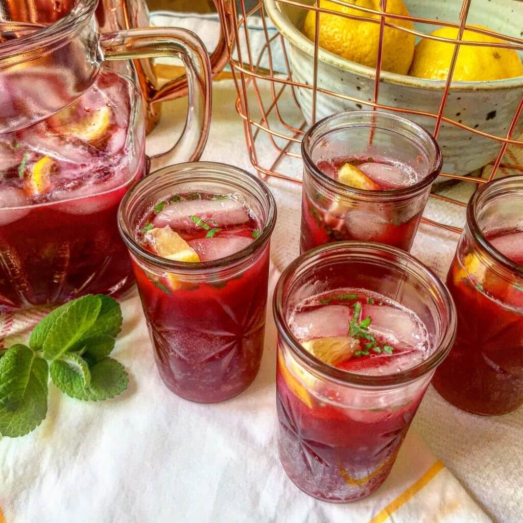 A glass pitcher and four glasses filled with a red beverage with ice, lemon slices, and mint, on a kitchen counter with lemons and mint leaves nearby.