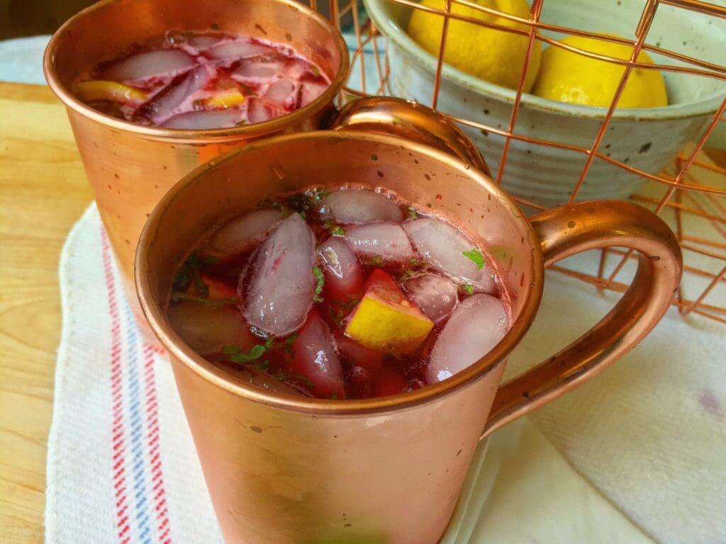 Two copper mugs filled with a red beverage, ice, lemon slices, and mint, on a kitchen counter with a bowl of lemons in the background.