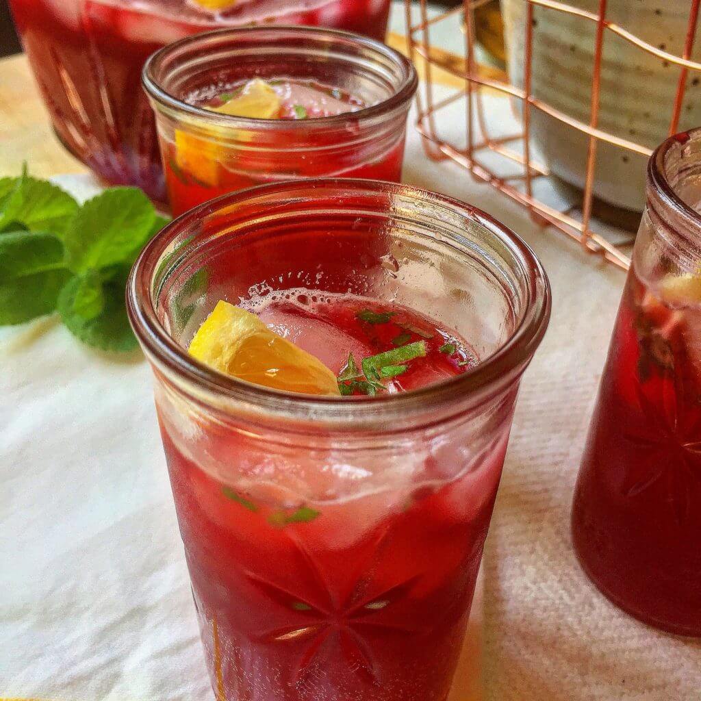 A glass filled with a red beverage, ice, lemon slices, and mint, with more glasses, mint leaves, and a pitcher in the background.
