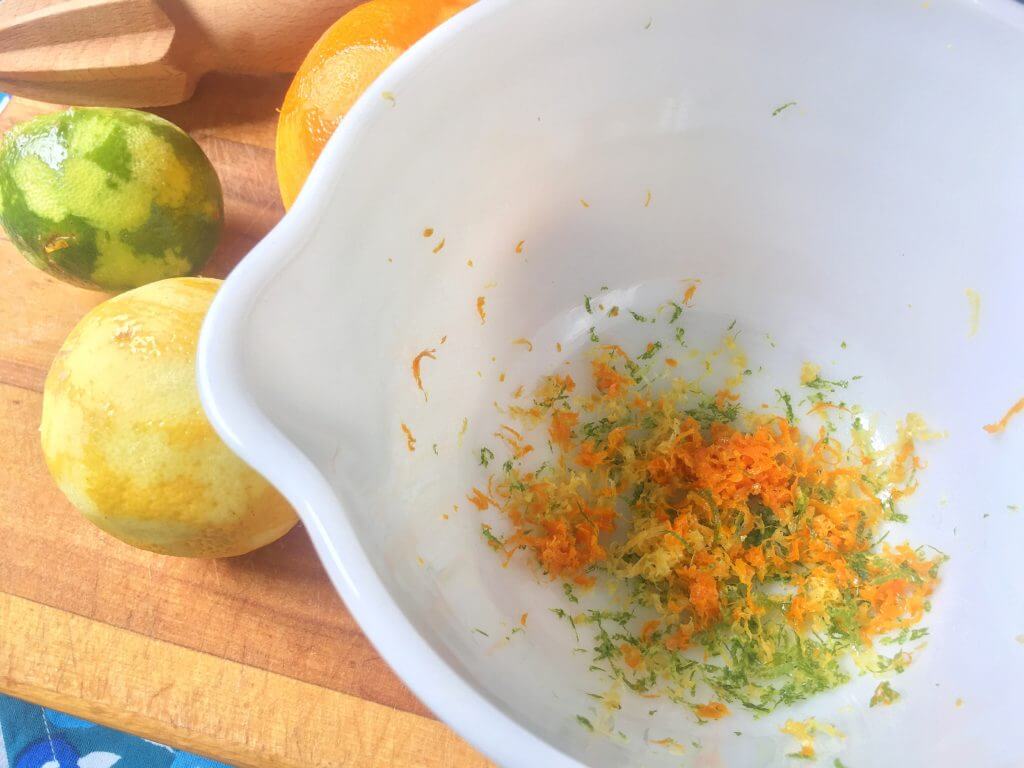 A white mixing bowl with citrus zest from an orange, lemon, and lime, placed on a wooden cutting board next to the peeled fruits and a wooden citrus reamer.