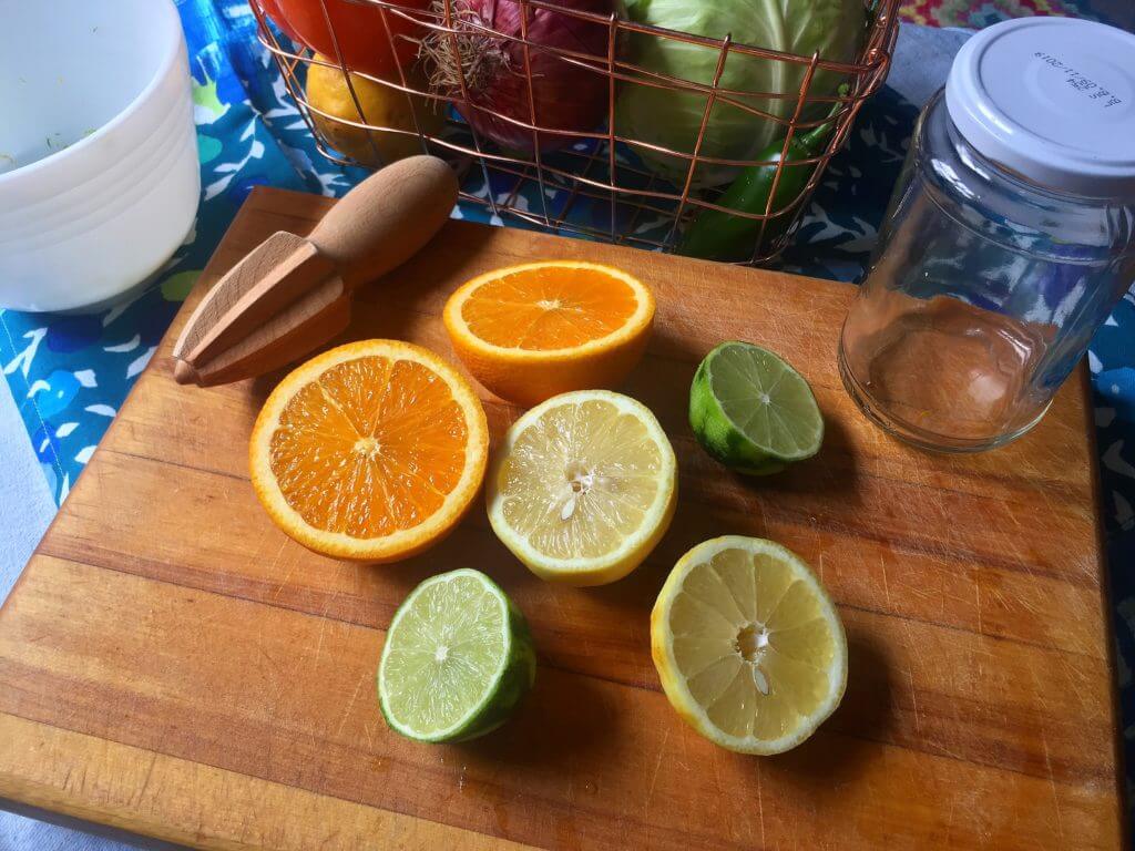 A wooden cutting board with halved citrus fruits (an orange, a lemon, and a lime), a wooden citrus reamer, an empty glass jar, and a wire basket with various vegetables in the background.