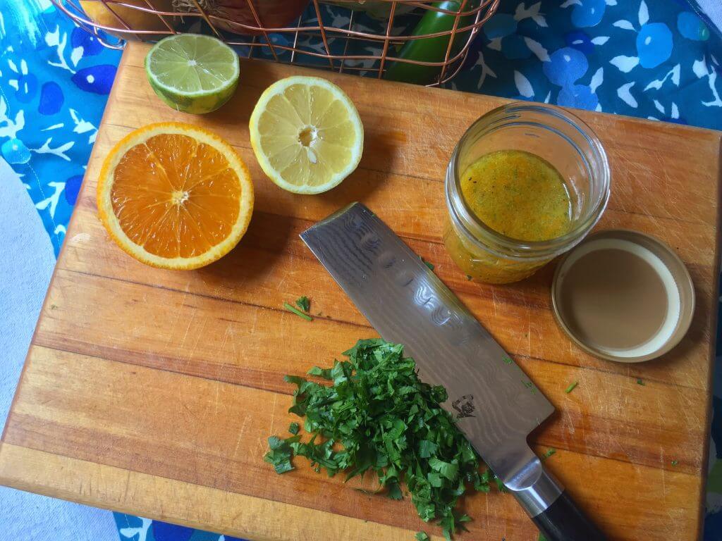A wooden cutting board with halved citrus fruits (an orange, a lemon, and a lime), a glass jar containing citrus dressing, a pile of chopped cilantro, and a knife, with a wire basket of vegetables in the background.