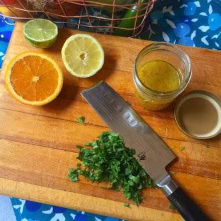 A wooden cutting board with halved citrus fruits (an orange, a lemon, and a lime), a glass jar containing citrus dressing, a pile of chopped cilantro, and a knife, with a wire basket of vegetables in the background.