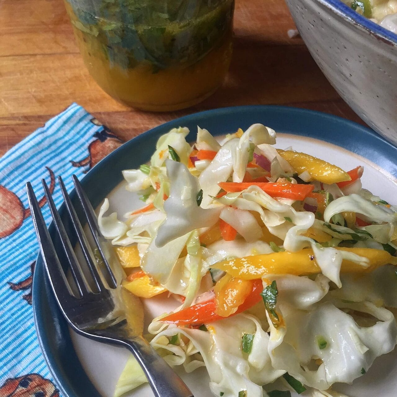 A plate of fresh coleslaw made with shredded cabbage, carrots, bell peppers, onions, and mango, served with a fork, with a jar of citrus dressing and a larger bowl of coleslaw in the background.