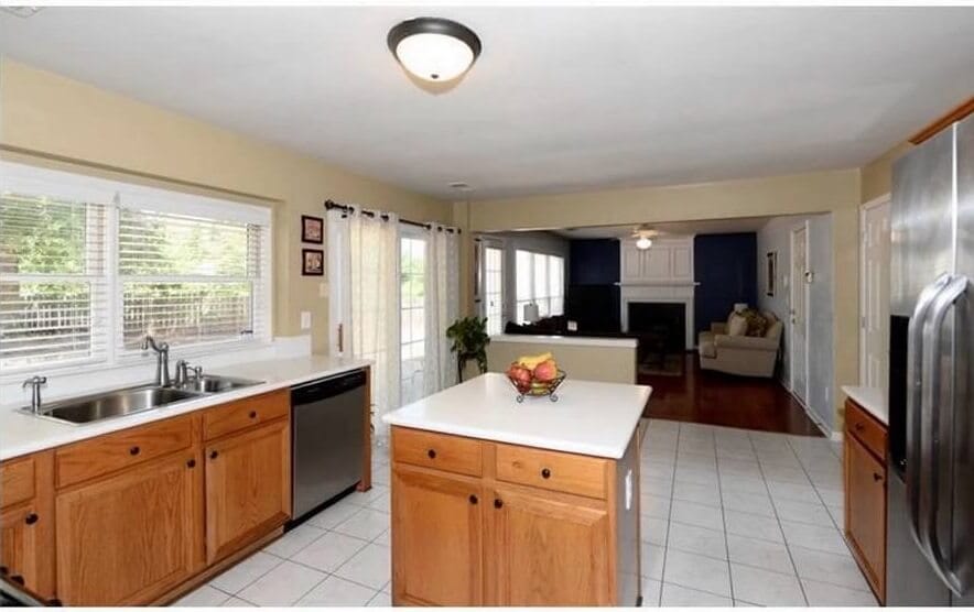 the before photo of a spacious kitchen with an island, wooden lower cabinets, white countertops, and stainless steel appliances.