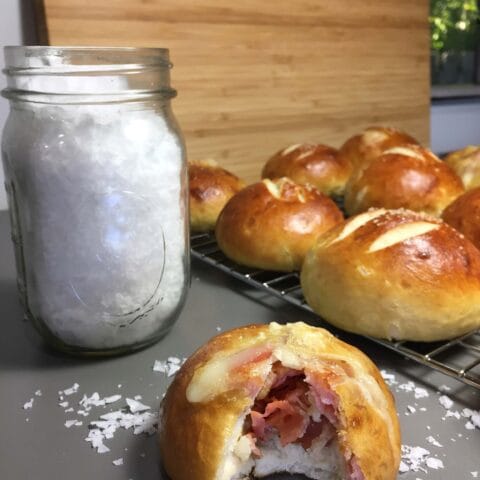 A jar of coarse salt next to a batch of freshly baked rolls, one of which has a bite taken out to reveal a bacon filling.