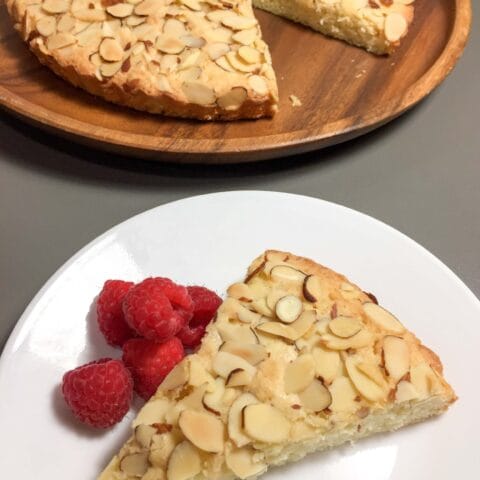 A slice of almond cake served with fresh raspberries on a white plate, with the remaining cake on a wooden tray in the background.