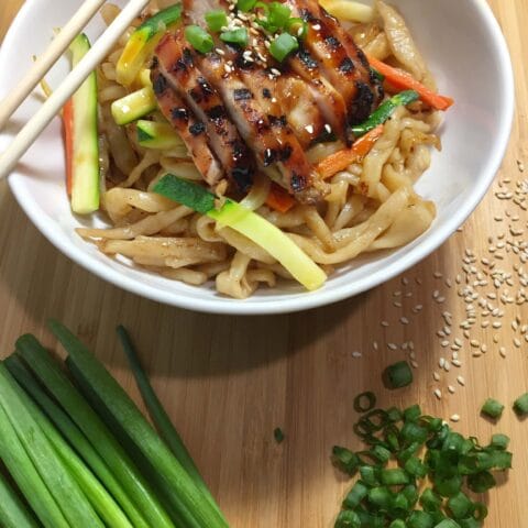 A bowl of noodles topped with sliced grilled chicken, julienned vegetables, green onions, and sesame seeds, with chopsticks resting on the bowl and green onions on the side.