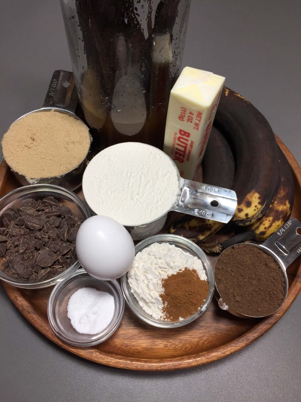 A wooden tray with baking ingredients: bananas, butter, brown sugar, chocolate chips, an egg, flour, cocoa powder, cinnamon, and salt.