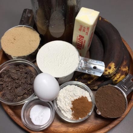 A wooden tray with baking ingredients: bananas, butter, brown sugar, chocolate chips, an egg, flour, cocoa powder, cinnamon, and salt.