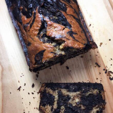 A loaf of marbled banana bread with one slice cut, showing the swirled pattern inside.