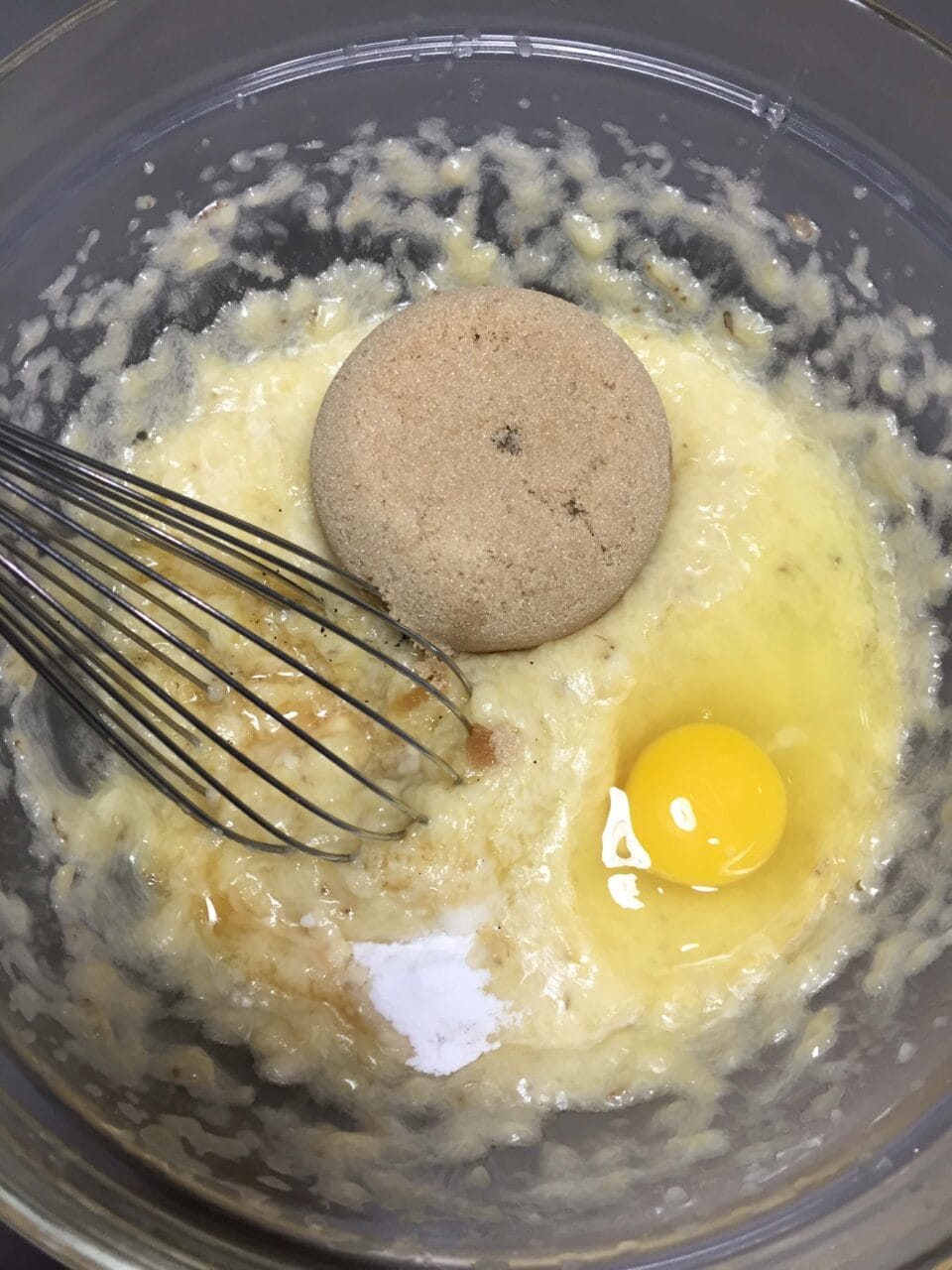 A mixing bowl with mashed bananas, an egg, brown sugar, and other ingredients being mixed with a whisk.