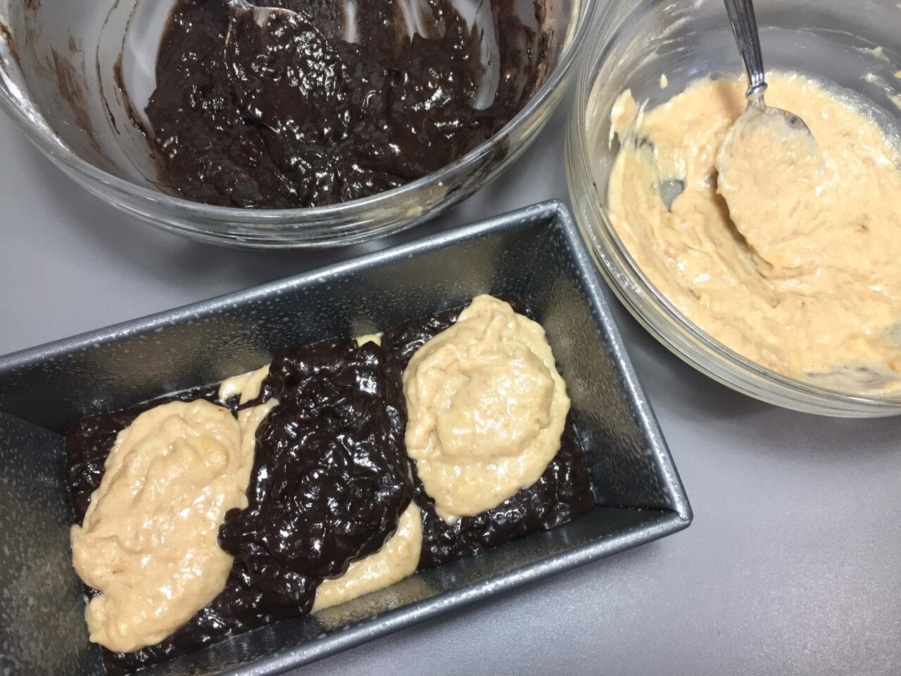 Two types of batter, one light and one dark, are being spooned into a loaf pan.