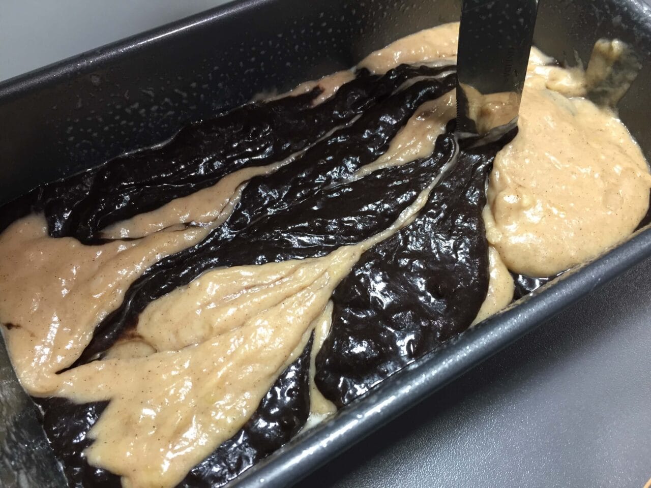A loaf pan with swirled light and dark batter being mixed with a knife.