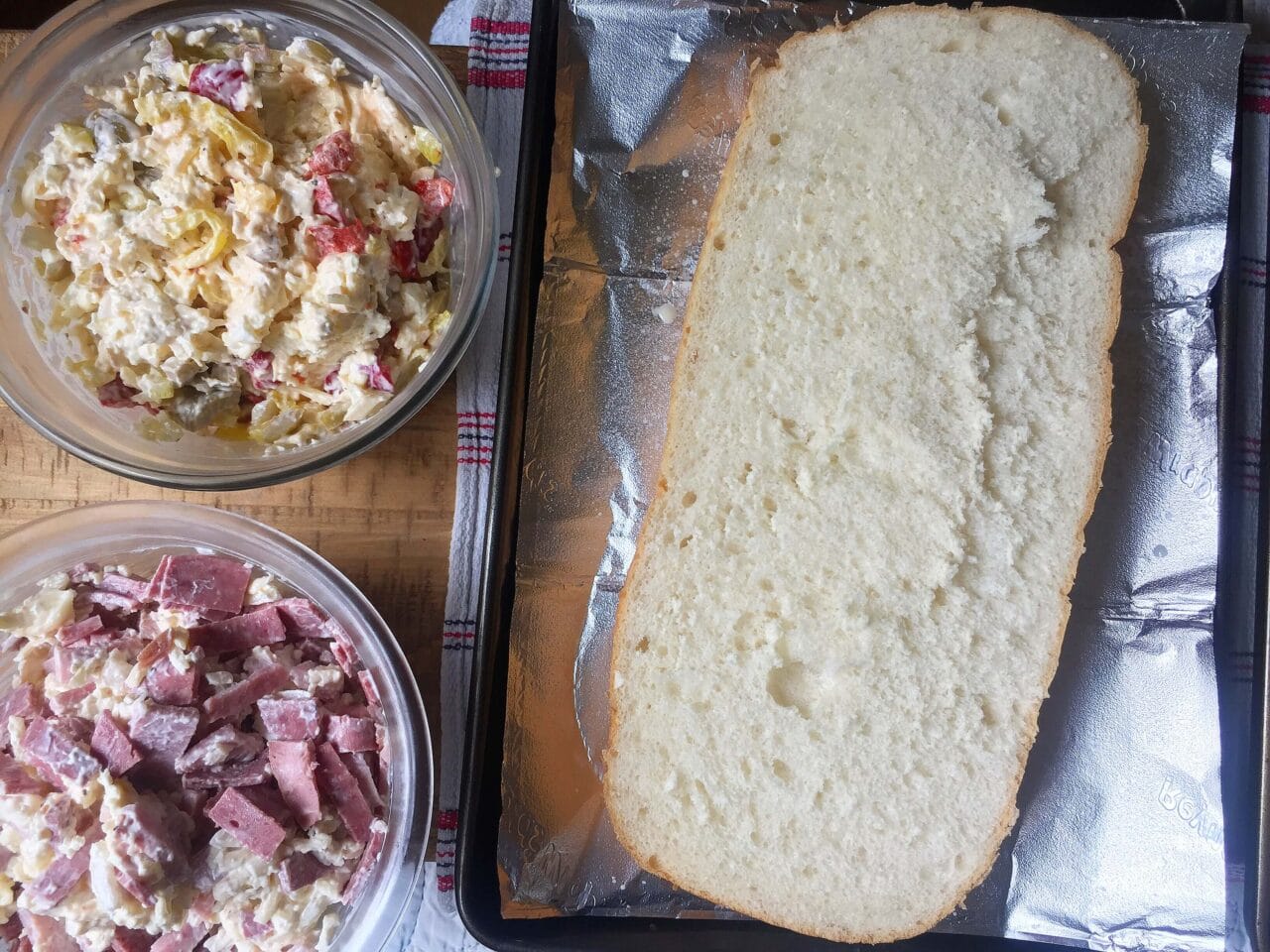 A loaf of bread on a baking sheet next to bowls of mixed cheese, meats, and peppers.