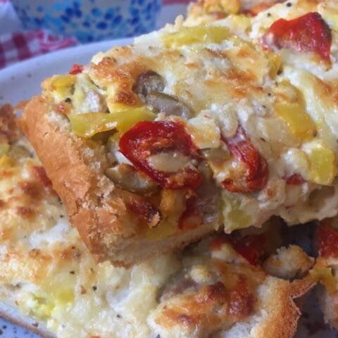 Slices of bread topped with melted cheese, peppers, and olives, with two drinks in the background.