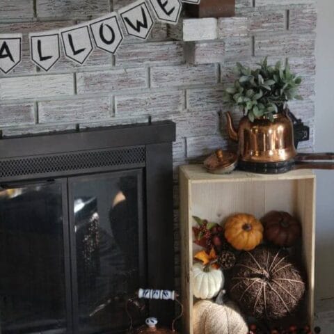 A fall fireplace display with a "Halloween" banner, a crate of pumpkins, and a copper kettle with greenery.
