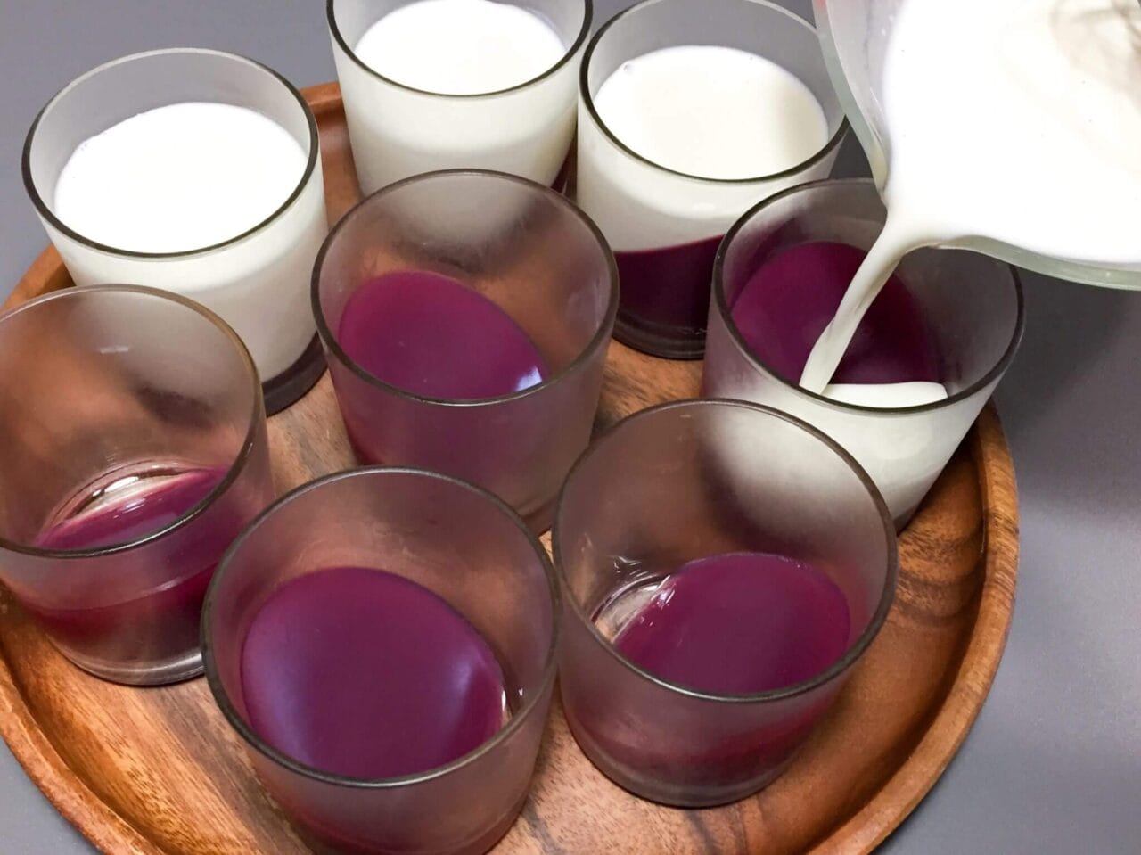 Several glass cups filled with red liquid being topped with a white liquid, all arranged on a wooden tray.
