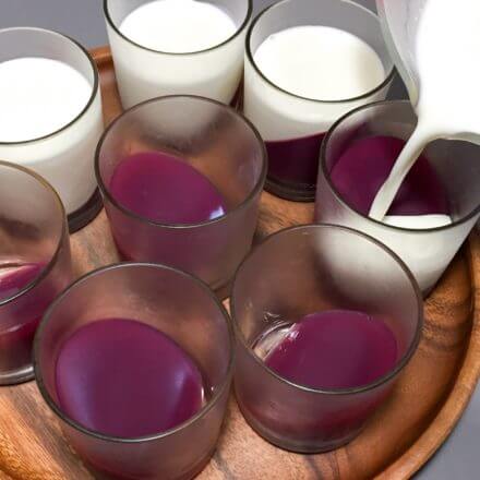 Several glass cups filled with red liquid being topped with a white liquid, all arranged on a wooden tray.