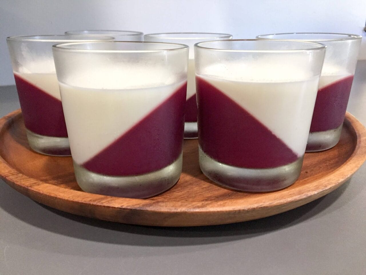 Several glass cups containing a two-layer dessert with a diagonal layer of red on the bottom and a white layer on top, with small red spots on the surface, arranged on a wooden tray.