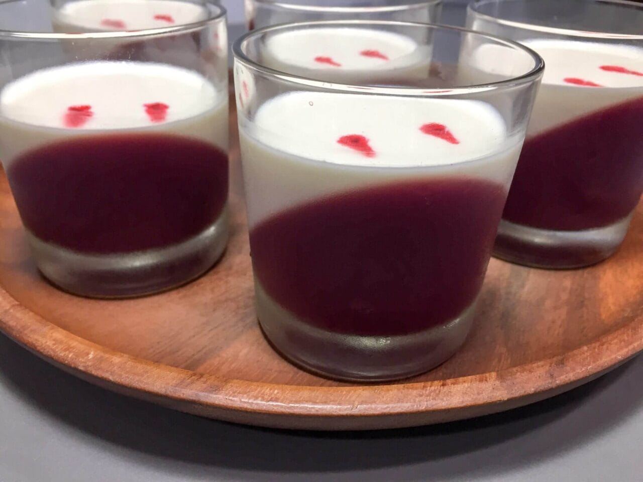 Several glass cups containing a two-layer dessert with a diagonal layer of red on the bottom and a white layer on top, with small red spots on the surface, arranged on a wooden tray with a pair of plastic vampire fangs.