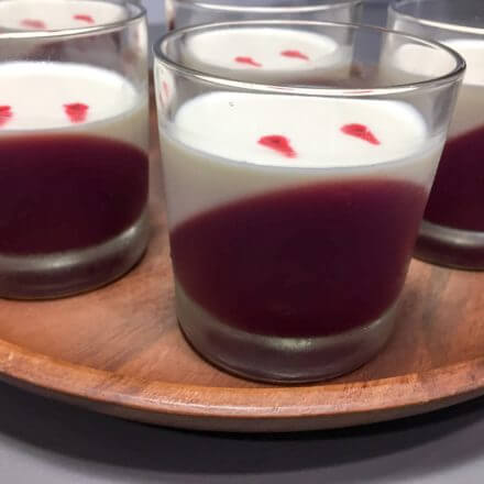 Several glass cups containing a two-layer dessert with a diagonal layer of red on the bottom and a white layer on top, with small red spots on the surface, arranged on a wooden tray.
