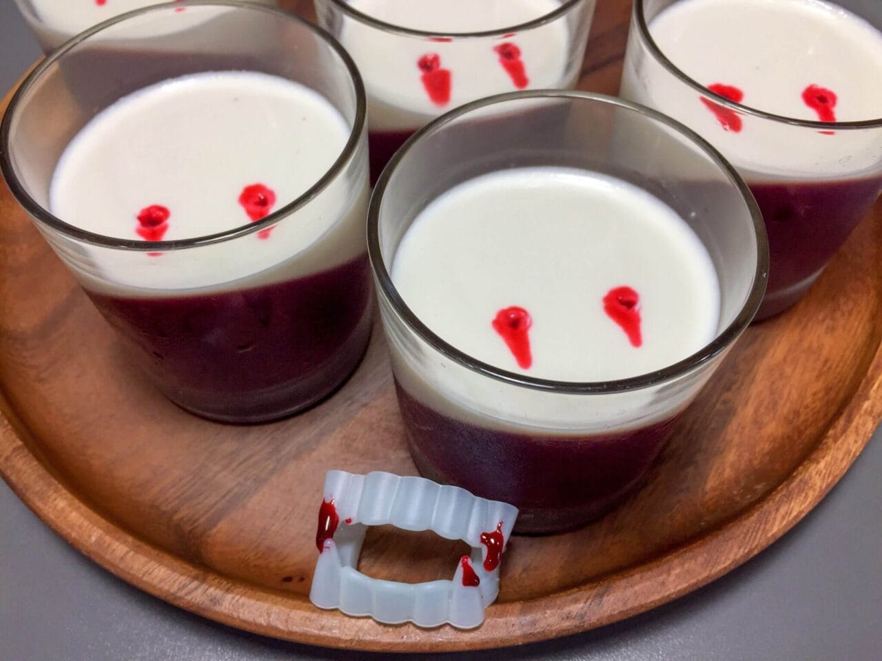 Several glass cups containing a two-layer dessert with a diagonal layer of red on the bottom and a white layer on top, with small red spots on the surface, arranged on a wooden tray with a pair of plastic vampire fangs.