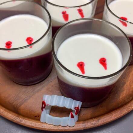 Several glass cups containing a two-layer dessert with a diagonal layer of red on the bottom and a white layer on top, with small red spots on the surface, arranged on a wooden tray with a pair of plastic vampire fangs.