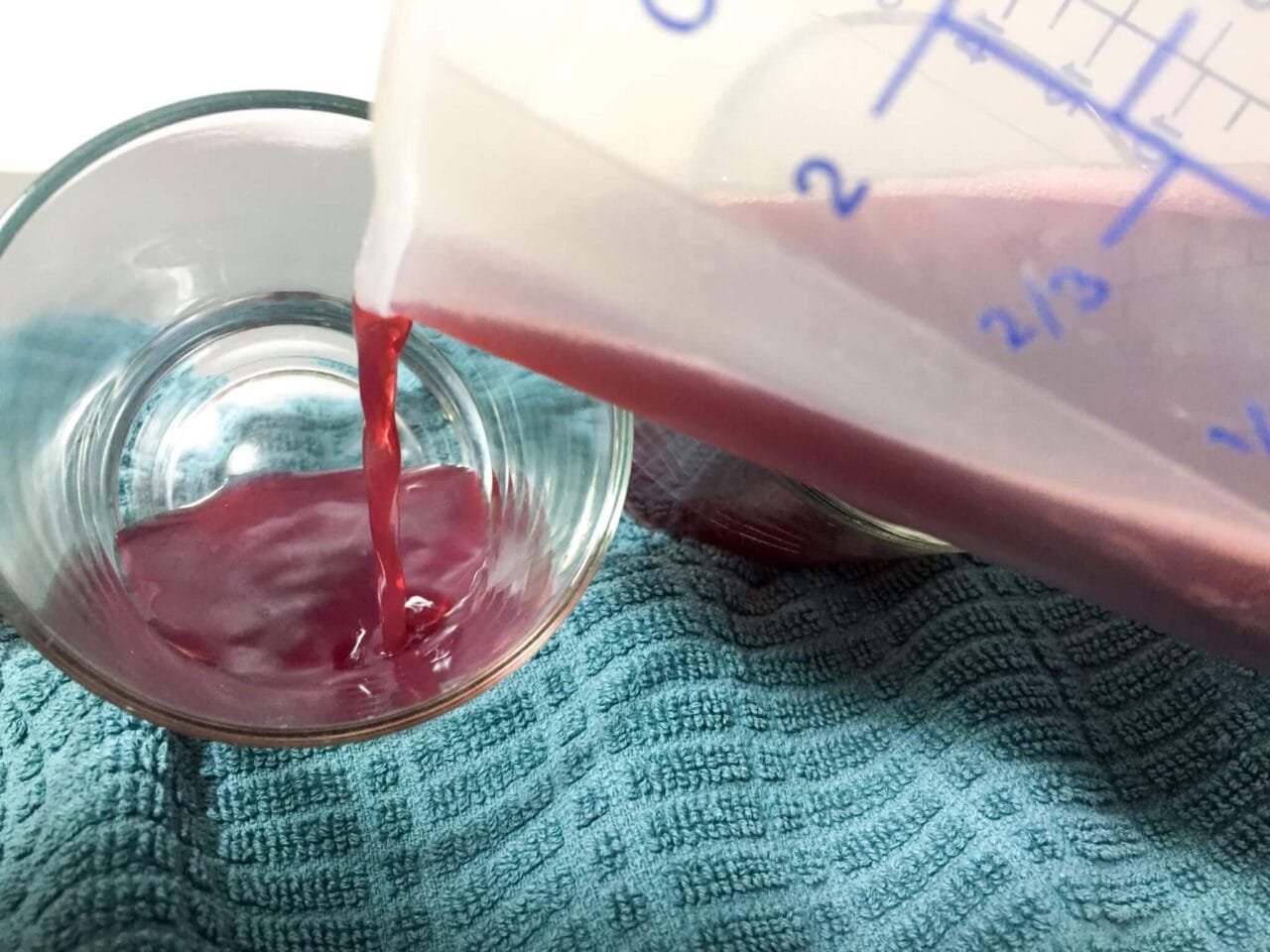 Red liquid being poured from a measuring cup into a glass bowl on a blue cloth.