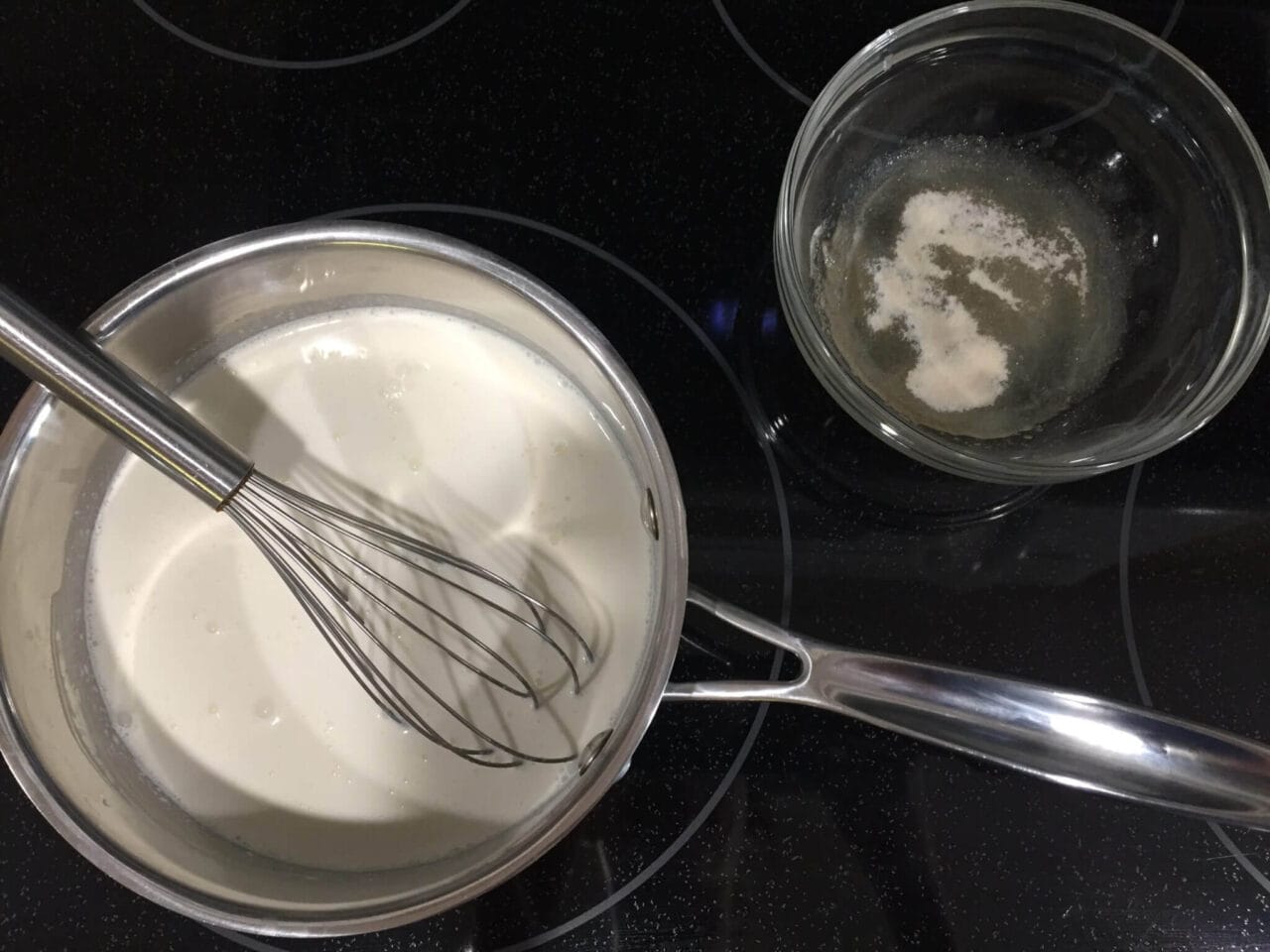 A saucepan with a whisk mixing a white liquid on a stovetop next to a glass bowl containing a gelatin mixture.