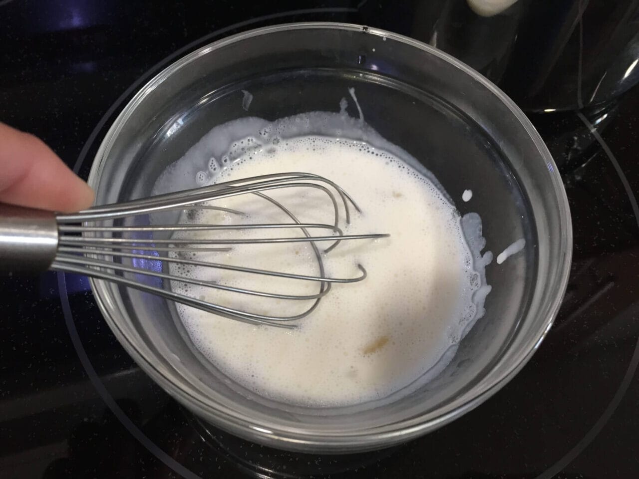 A whisk being used to mix a white liquid in a glass bowl.