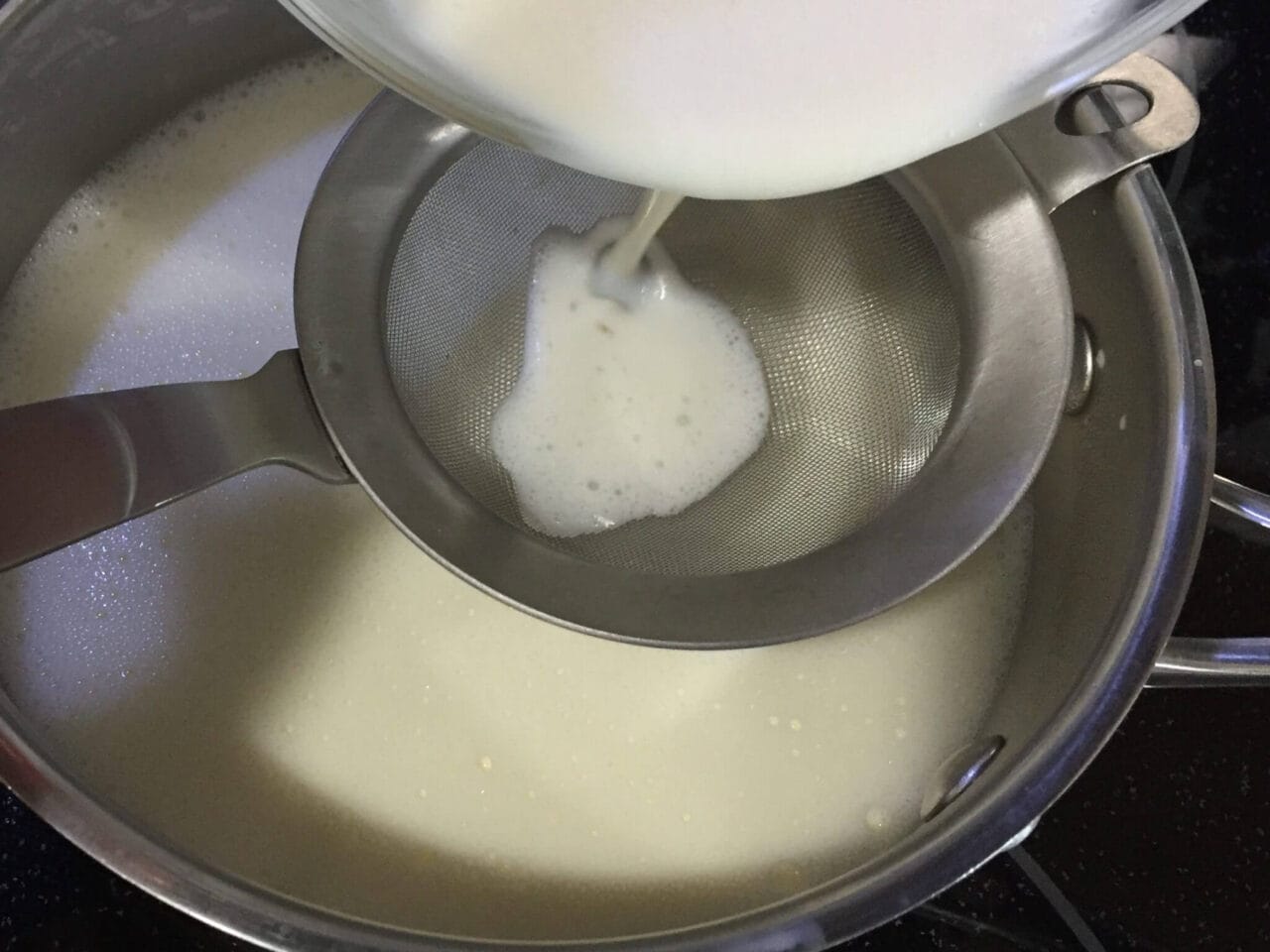 A white liquid being poured through a fine mesh strainer into a saucepan.