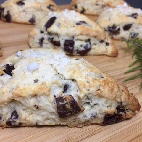 Freshly baked scones with chocolate chunks are arranged on a wooden surface next to a sprig of rosemary.