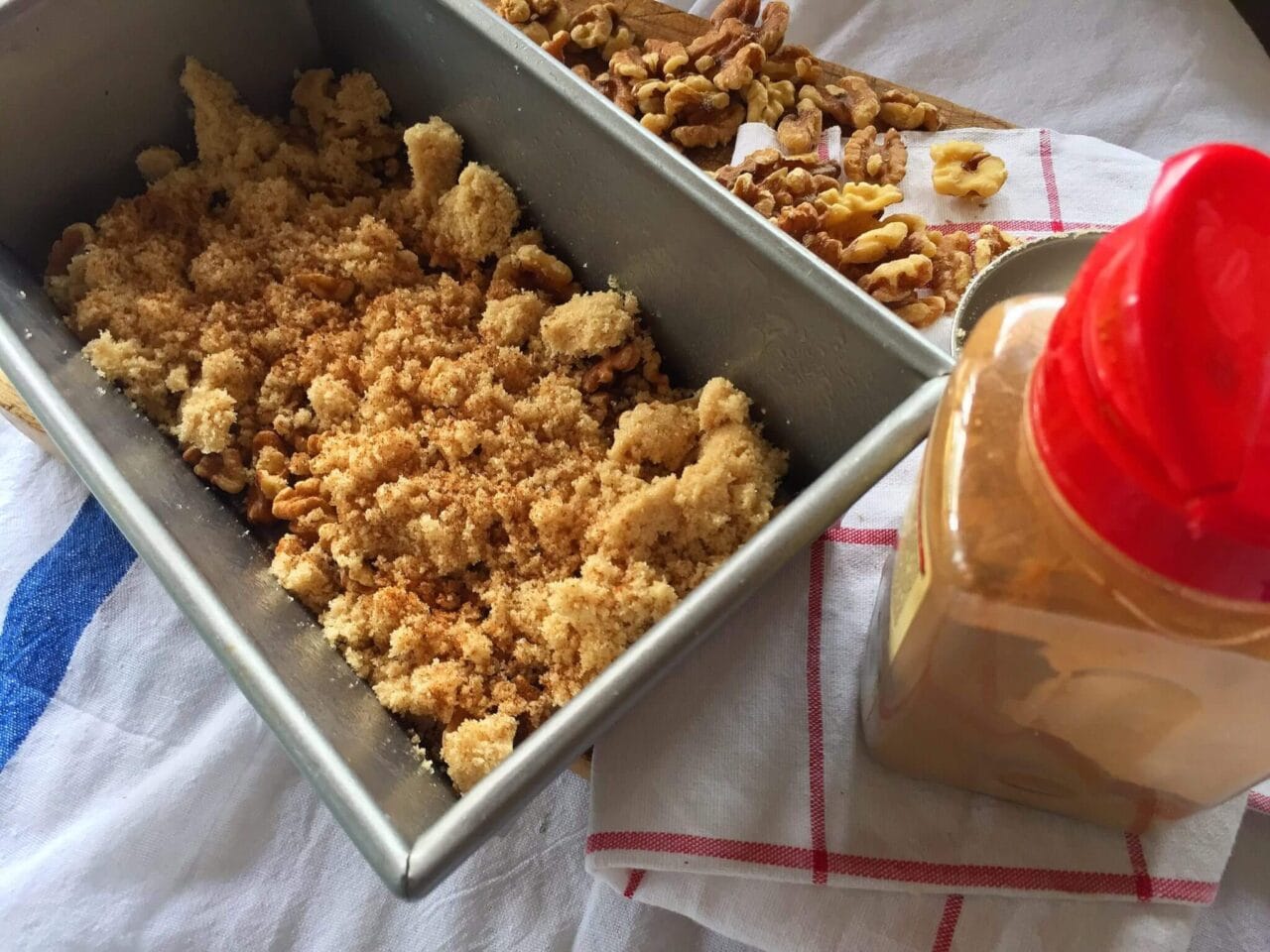 A loaf pan filled with a crumbly mixture, next to a container of cinnamon and scattered walnuts.