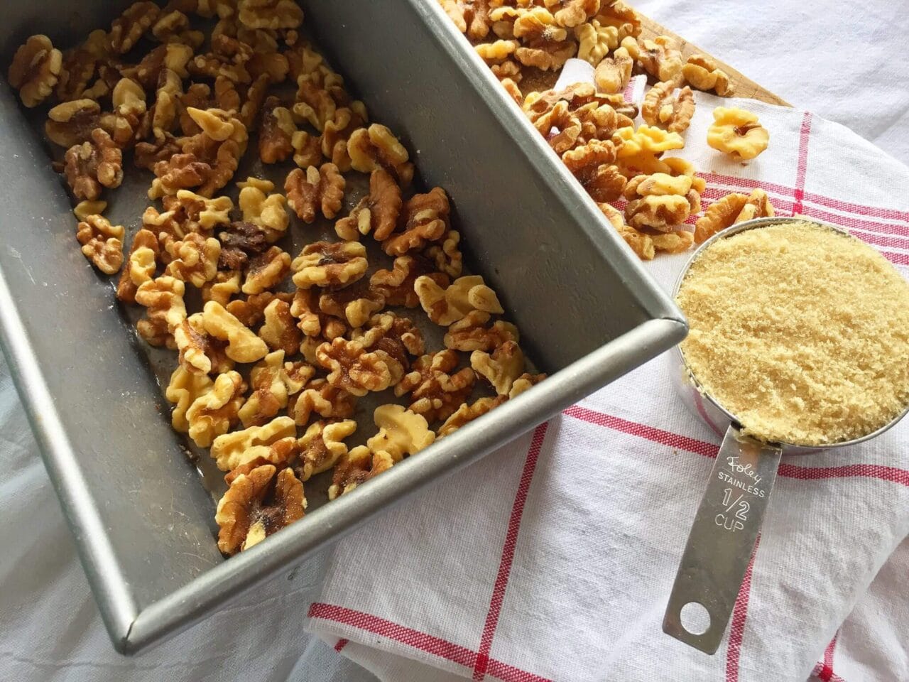 A loaf pan with walnuts at the bottom, next to a half-cup measuring cup filled with brown sugar.