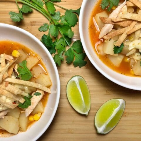 Two bowls of tortilla soup with shredded chicken, diced potatoes, corn, and topped with crispy tortilla strips and cilantro, with lime wedges and cilantro sprigs on the side.